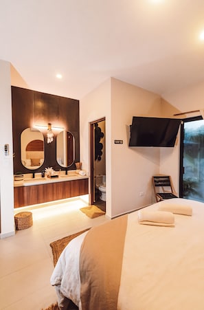 A modern hotel room with a large bed, a vanity with a mirror, a flat-screen TV mounted on the wall, and a window offering a view of the outdoors.