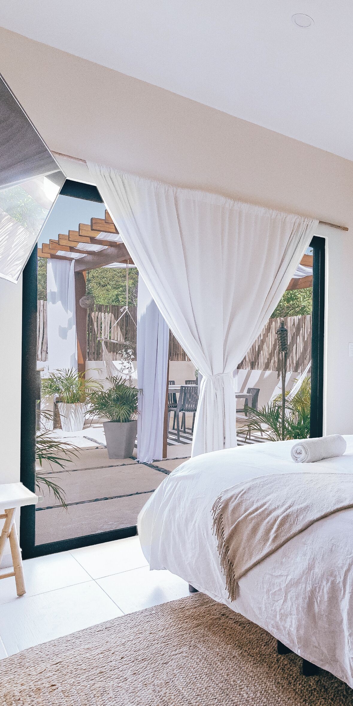 Een slaapkamer met een groot bed, uitzicht op een terras buiten met stoelen en planten, en een houten pergola.