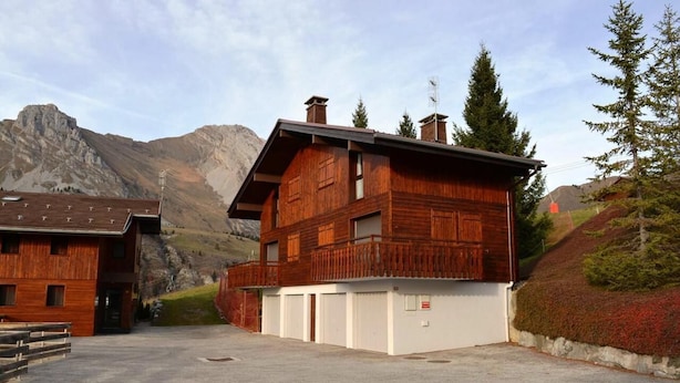 En træchalet med balkon, garage og et bjergrigt landskab i baggrunden.