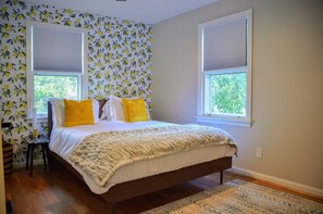 A bedroom with a bed, two windows with blinds, a patterned wall, and a wooden floor.