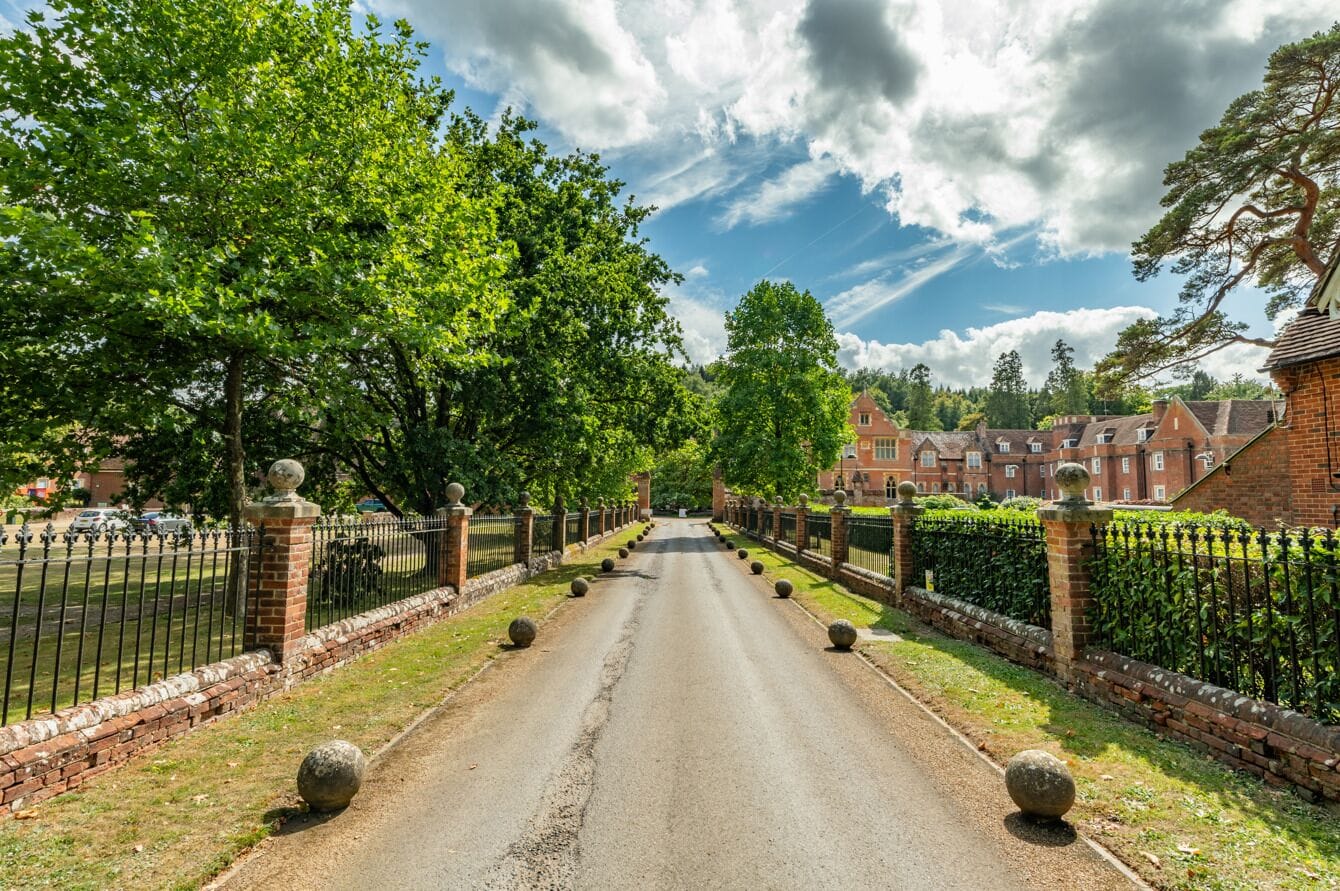A paved pathway flanked by brick walls and metal fencing, leading to a building with multiple windows and a traditional architectural style.