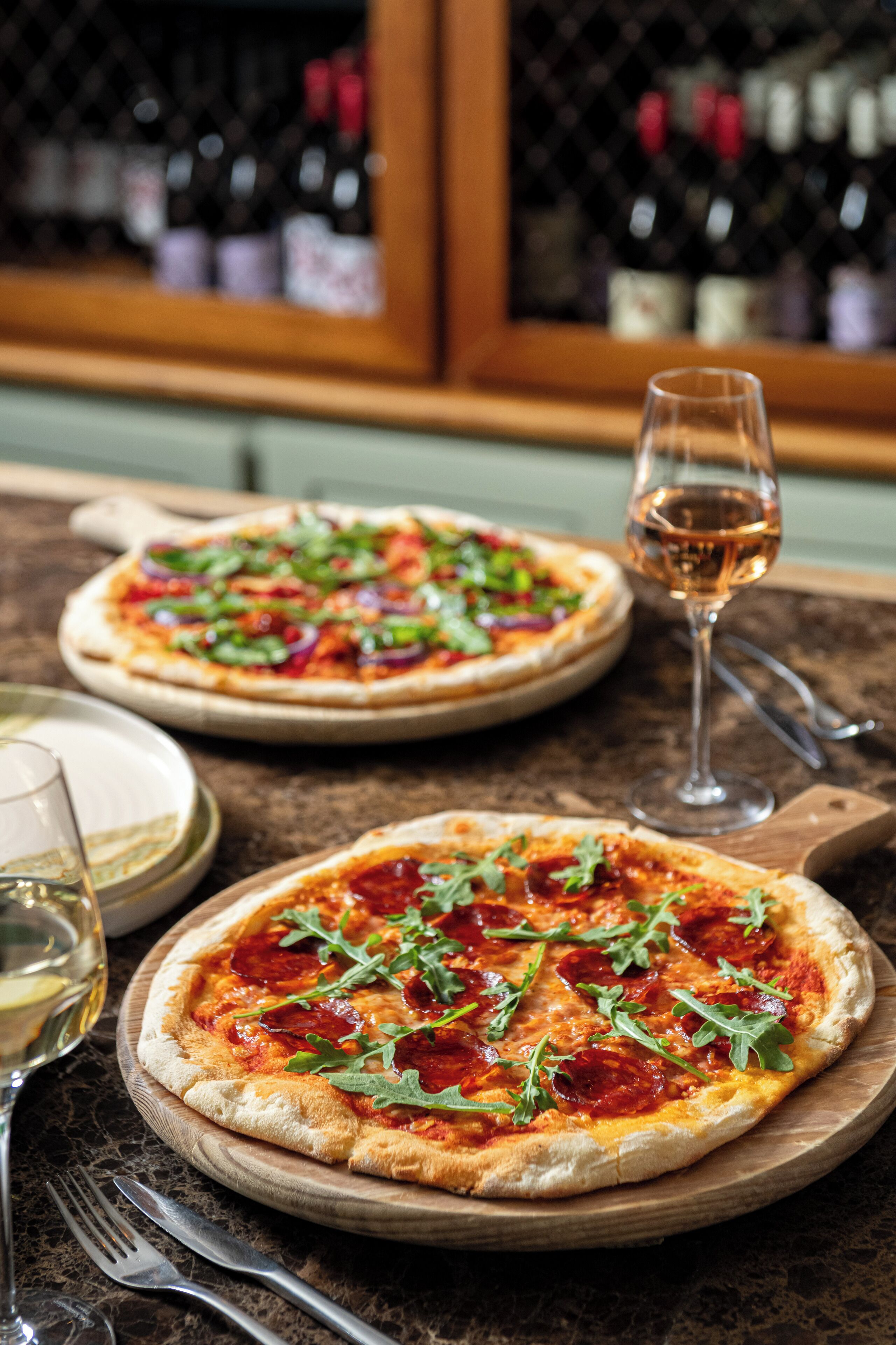 Two pizzas on wooden boards with a wine glass and cutlery beside them.