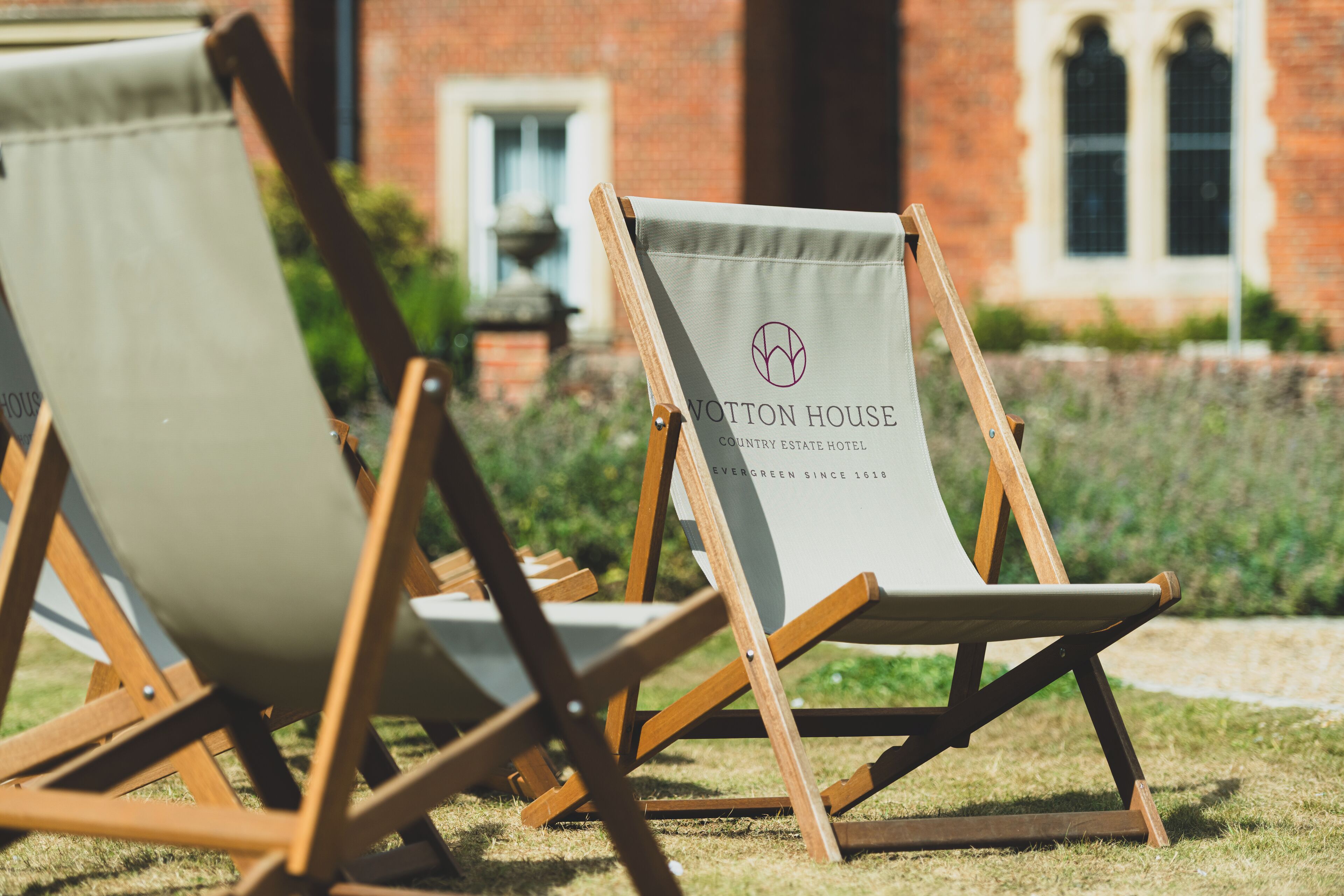 Two canvas lounge chairs with 'WOTTON HOUSE COUNTRY ESTATE HOTEL' branding, positioned on grass in front of a brick building.