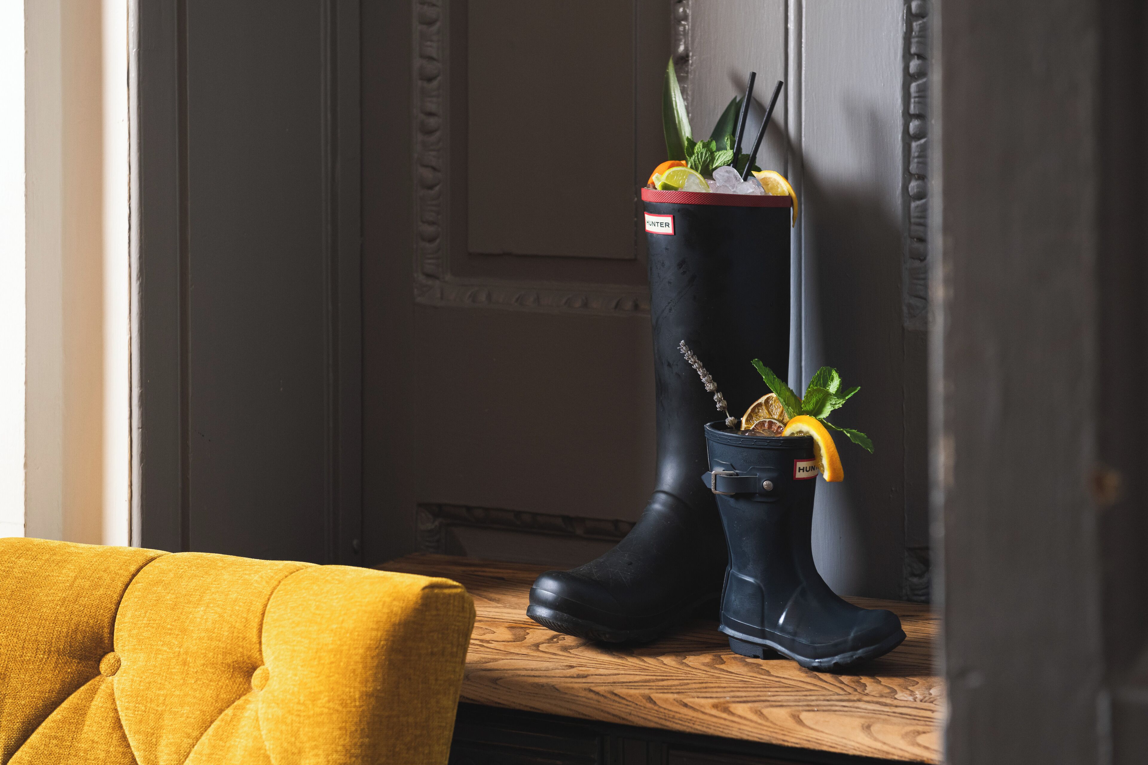 A pair of black rubber boots with a red tag on the side, placed on a wooden surface next to a yellow chair.