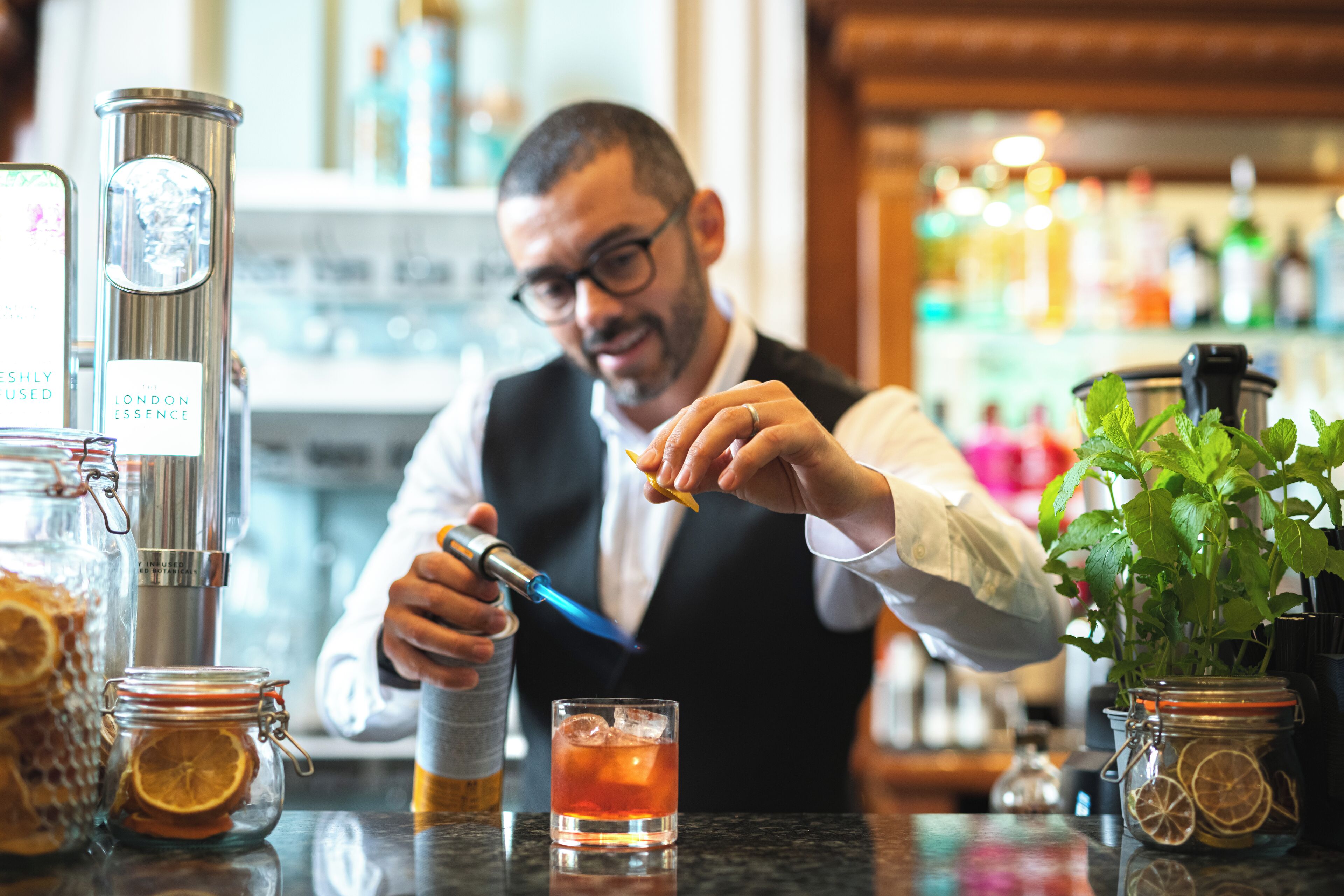 Un cantinero preparando una bebida, con énfasis en la zona del bar y una planta en maceta.