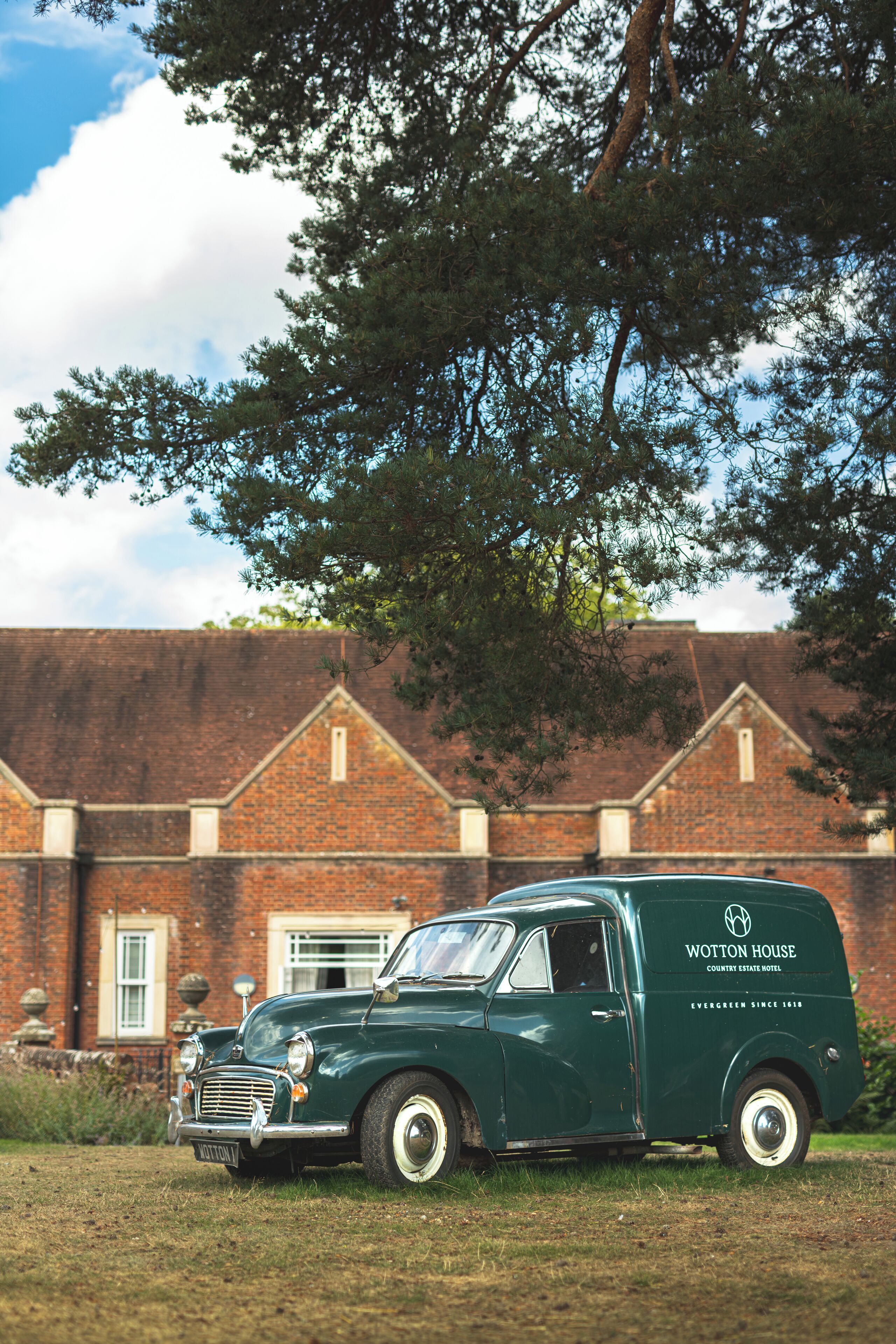 Un vieux camion vert est garé devant un bâtiment en briques, et un grand arbre se dresse au-dessus.