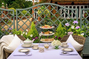 Un juego de servicio para té de tres niveles sobre una mesa, con una silla de mimbre y una verja metálica verde al fondo.