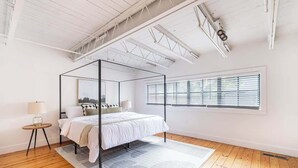 Een moderne slaapkamer met een hemelbed, een groot raam met zonwering, en een wit plafond met zichtbare balken.