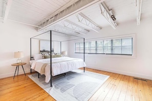 Ein modernes Schlafzimmer mit einem Himmelbett, einem großen Fenster mit Jalousien und einer weißen Decke mit freiliegenden Balken.