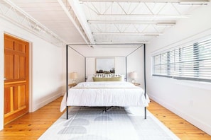 A bedroom with a four-poster bed, wooden flooring, a large window with blinds, and a white ceiling with exposed beams.
