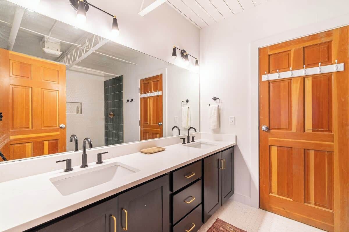 Une salle de bain avec un grand miroir, deux lavabos, des meubles sombres et une porte en bois.