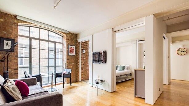 Una sala moderna con un ventanal grande, chimenea, zona de comedor y un dormitorio con cama y mesita de noche.