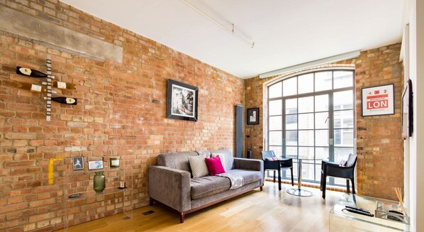 A room with a brick wall, a grey sofa, a glass table, and a large window. - Central Soho Lofts with Lift,  AirCon & Concierge (Greater London)