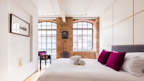 Un dormitorio con una cama grande, dos ventanas, una pared de ladrillo y un cuadro en la pared.