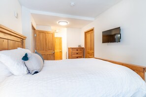 A bedroom with a large bed, wooden wardrobe, a television mounted on the wall, and a small table with a lamp. - 3 Minute Walk to Gondola w/ hot tub, pool, sauna! (Keystone)