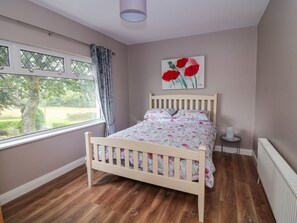 A bedroom with a wooden bed, a window with curtains, a radiator, and a painting of red flowers on the wall. - Rockwood Cottage (Castledockrell)