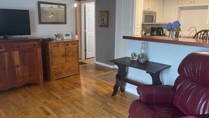 A living room with a wooden floor, a red leather chair, a wooden cabinet, a television, and a kitchen area with white cabinets and a microwave. - Cozy, Clean, and Convenient (Parsons)