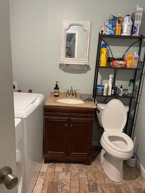 Een badkamer met een wit toilet, een houten wastafelmeubel met een wastafel, een spiegel boven het meubel en een plank met diverse toiletartikelen.