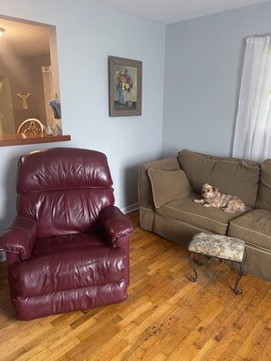 Een woonkamer met een houten vloer, een bruine bank, een rode lederen fauteuil en een klein krukje.