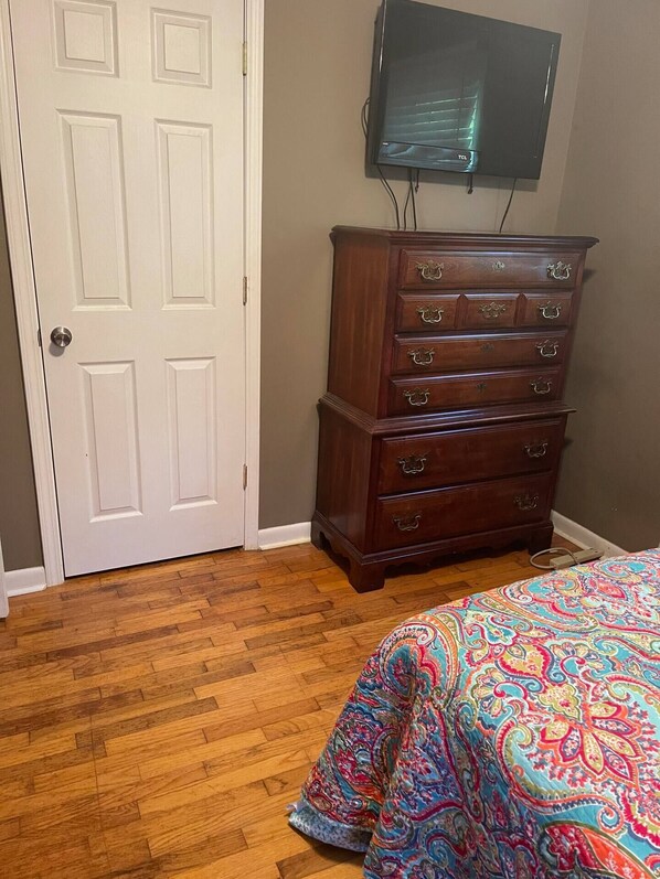 A bedroom with a wooden dresser, a bed with a colorful bedspread, a white door, and a flat-screen TV mounted on the wall. - Cozy, Clean, and Convenient (Parsons)