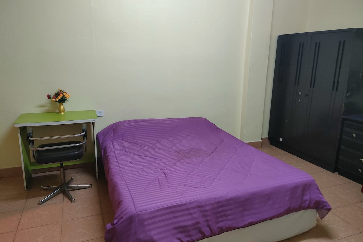A room with a bed, a desk with a chair, a wardrobe, and a vase with flowers.