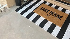 Un paillasson noir et blanc à rayures, sur lequel est inscrit le mot « LAKE HOUSE », une plante en pot et une porte avec un panneau en verre.