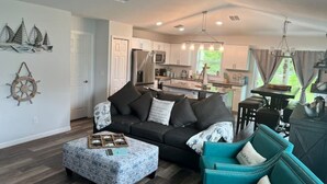 A modern living room with a dark grey sofa, teal armchairs, and a patterned ottoman. A ceiling fan and pendant lights are visible.