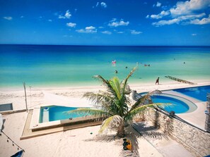 A beach with a pool, palm trees, and a clear blue sky.