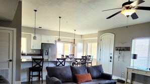 A modern living room with a grey sofa, a dining area with a table and chairs, a kitchen with a refrigerator, and a ceiling fan. - Beautiful New 2 Bedroom Condo in the heart of St. George (St. George)