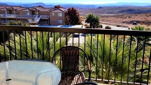 A balcony with a glass table and wicker chairs, overlooking a desert landscape with distant mountains and a residential building. - Beautiful New 2 Bedroom Condo in the heart of St. George (St. George)