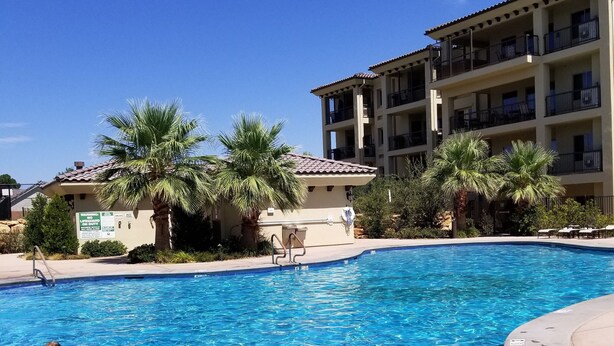 A swimming pool with clear blue water, surrounded by palm trees and a multi-story building with balconies. - Beautiful New 2 Bedroom Condo in the heart of St. George (St. George)