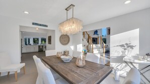 Un espace repas moderne avec une table en bois, des chaises blanches et un lustre. On aperçoit une cuisine au fond, et une pancarte indique « Beach House ».