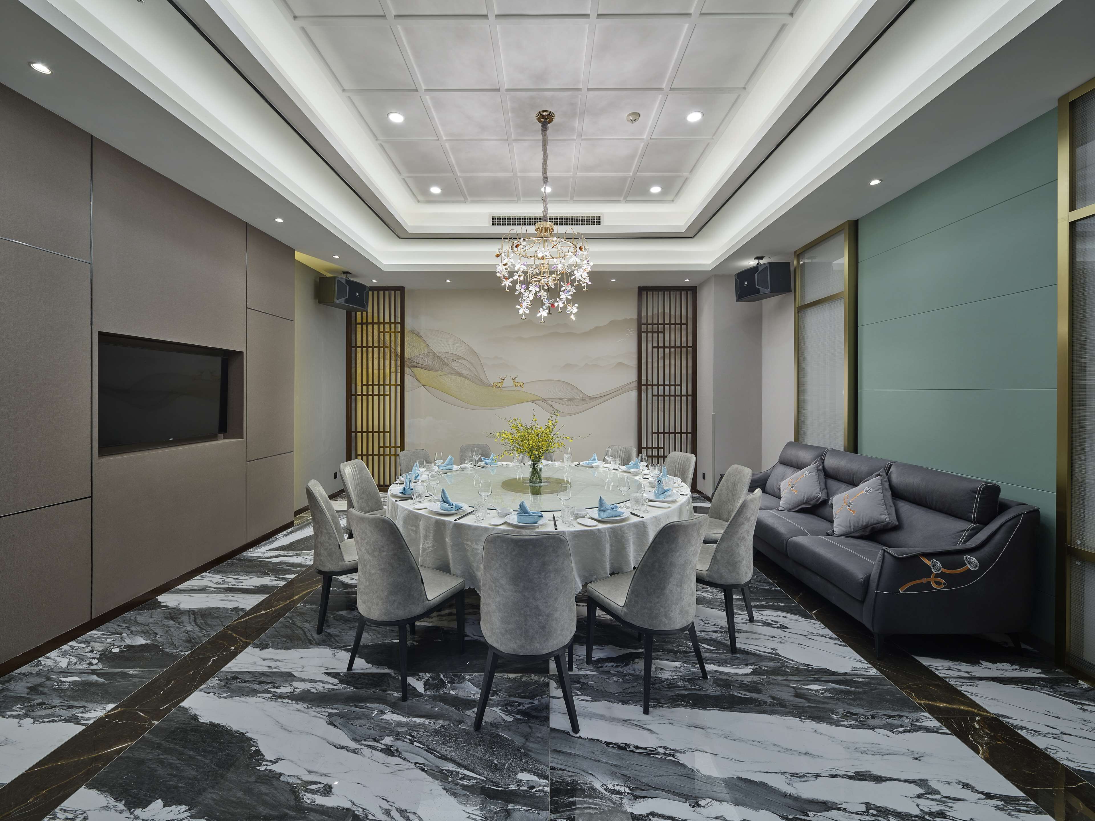 A modern conference room with a round table, grey upholstered chairs, a chandelier, and a grey sofa with cushions.