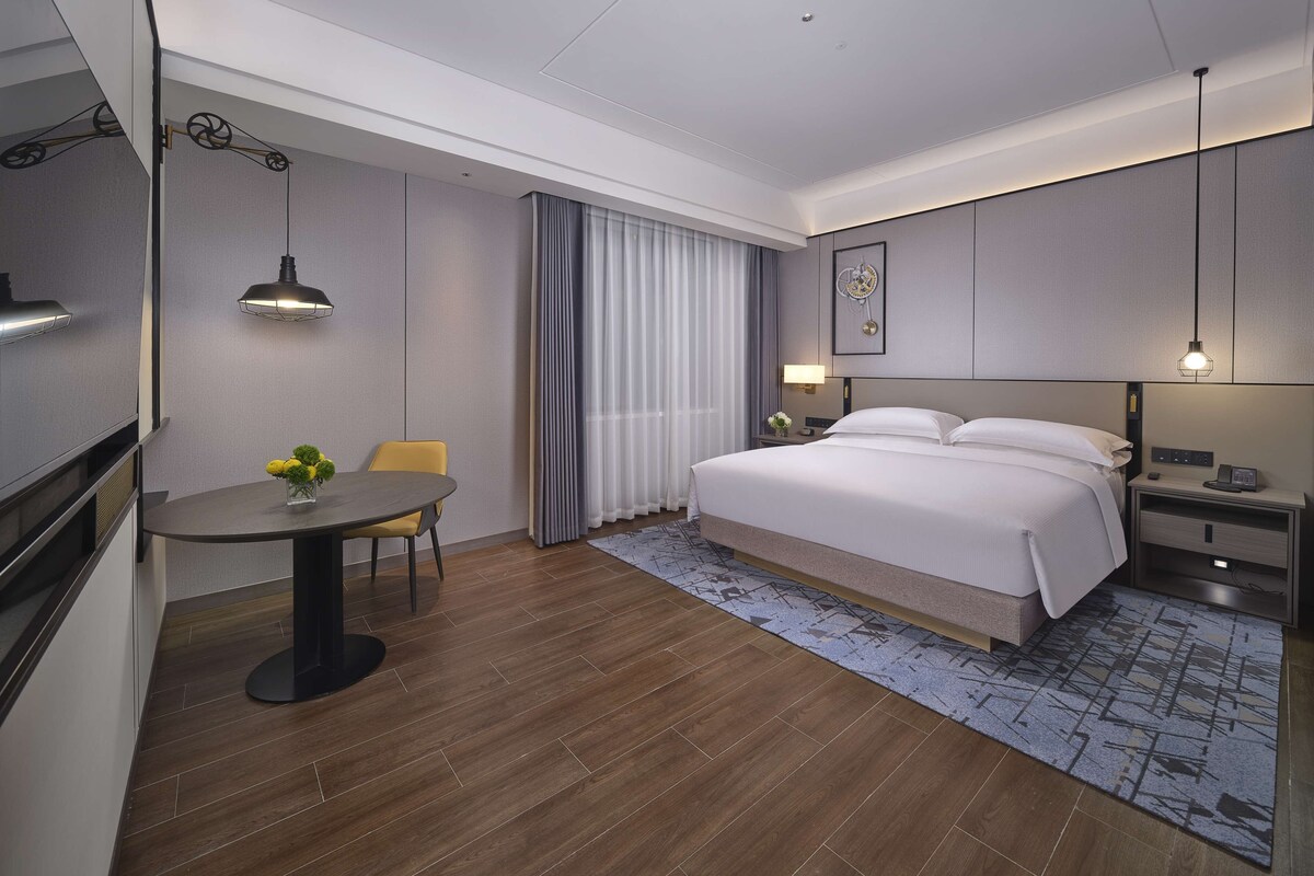 A modern hotel room with a bed, bedside table, lamp, and a dining area with a table and chairs.