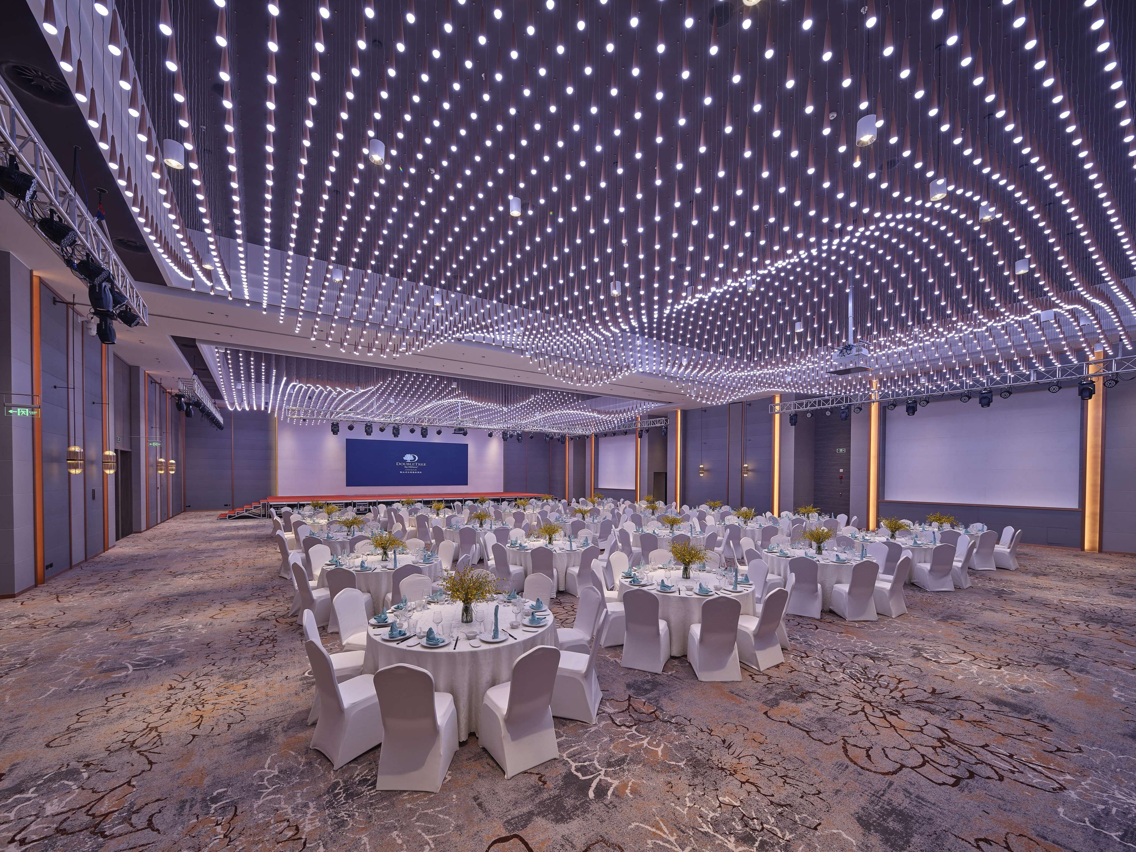 A spacious banquet hall with round tables set for events, featuring a modern ceiling design with hanging lights and a large screen at the front.