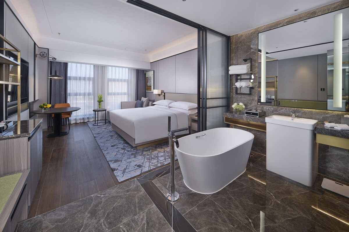A modern hotel room with a large bed, a separate bathroom with a bathtub, a dining area, and a large mirror.