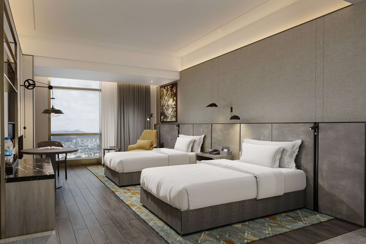 A modern hotel room with two beds, a desk, a chair, and a large window offering a city view.