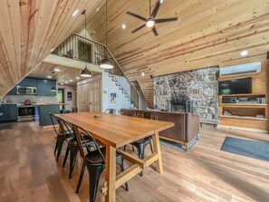 A spacious dining area with a wooden table and chairs, a kitchen with modern appliances, a stone fireplace, and a loft-style staircase.