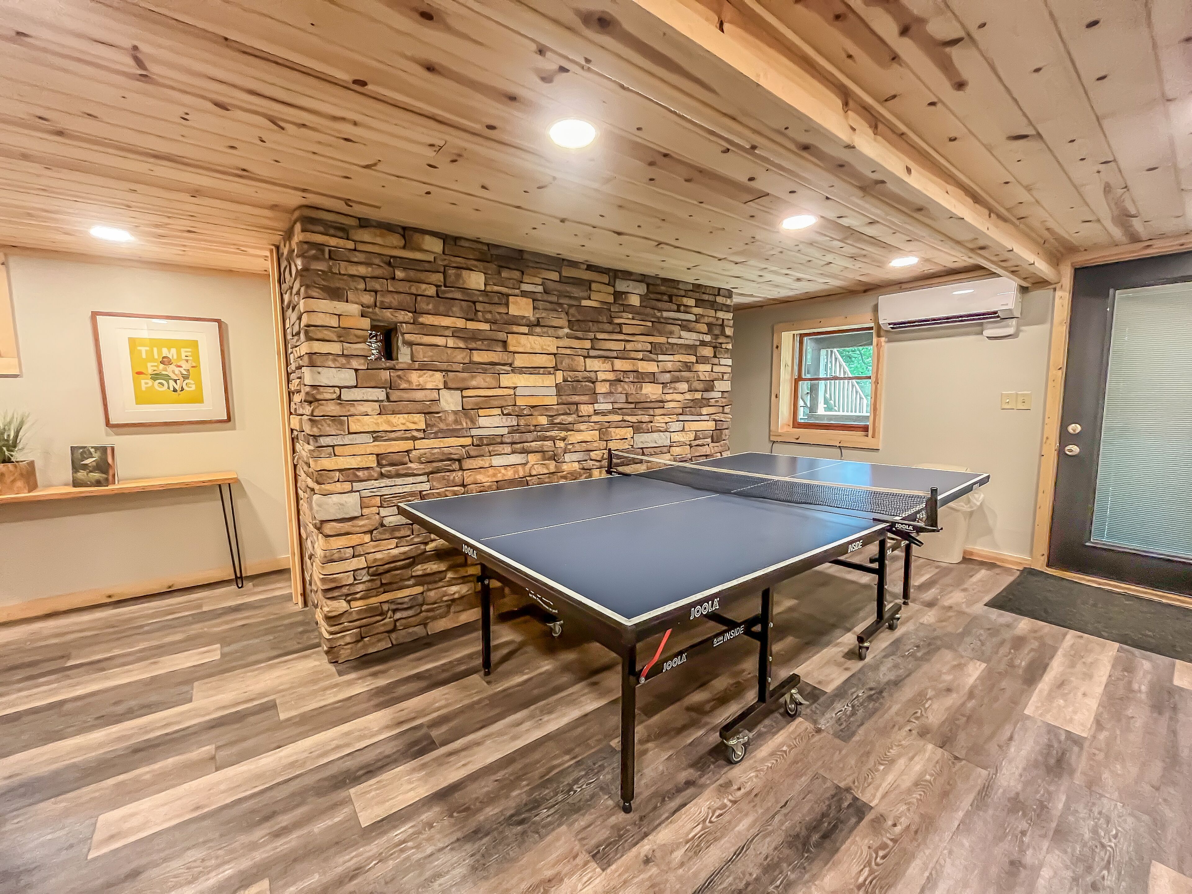 Una mesa de ping pong en una habitación con suelo de madera, pared de piedra y una puerta con ventana.