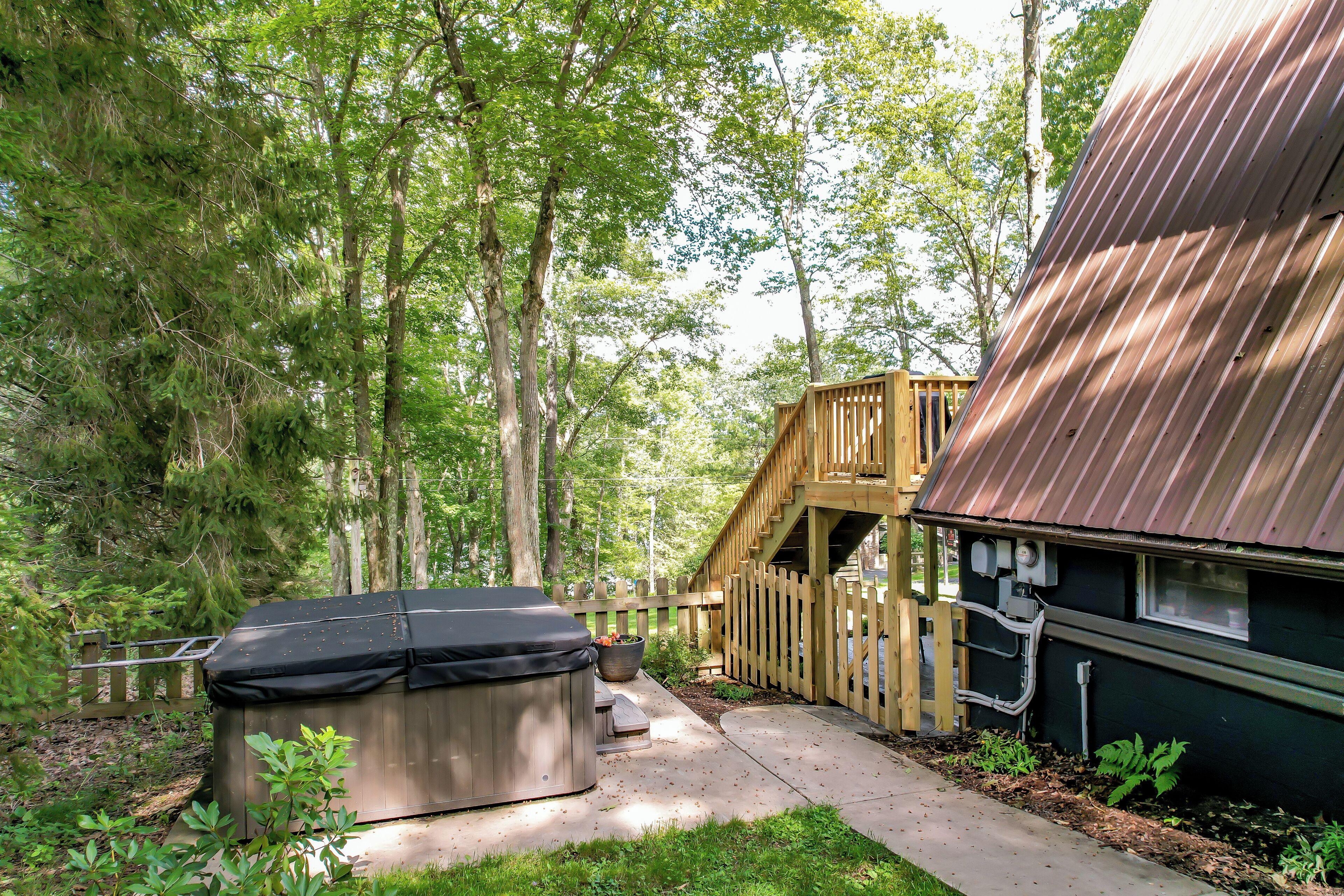 A wooden deck with a hot tub, surrounded by a forested area and a house with a metal roof.