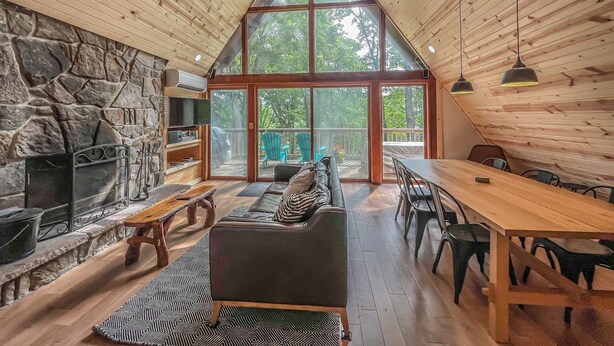 A cozy cabin interior with a stone fireplace, wooden ceiling, and large windows offering a view of the outdoors.