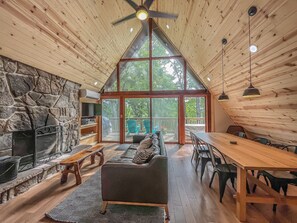 A cozy cabin interior with a stone fireplace, wooden ceiling, and large windows offering a view of the outdoors.