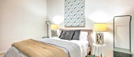 A neatly arranged bedroom with a bed, two bedside tables with lamps, a green chair, and a patterned wall hanging.