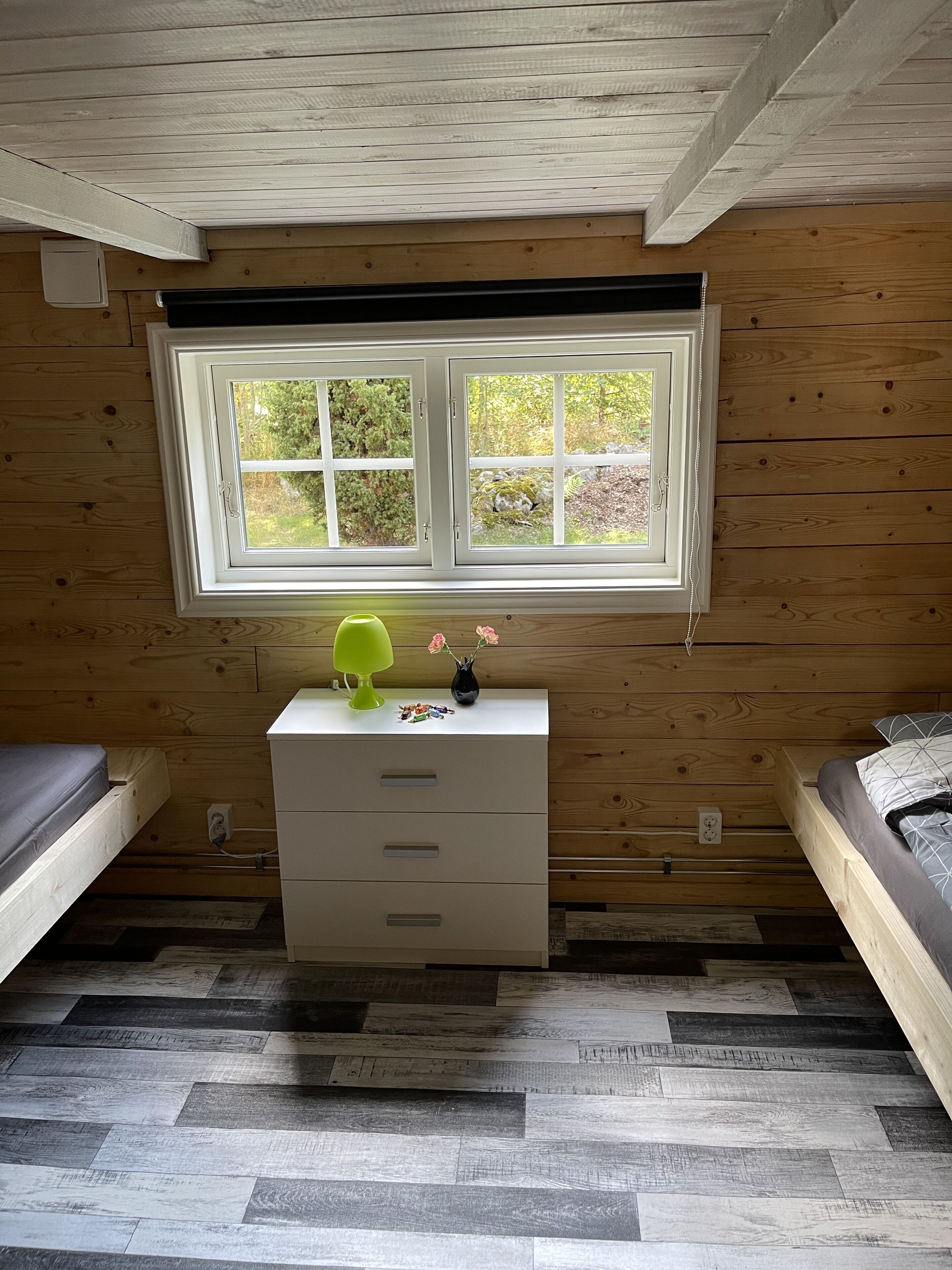 A room with a wooden ceiling, a window with a view of greenery, a white dresser, and a bed with gray bedding.