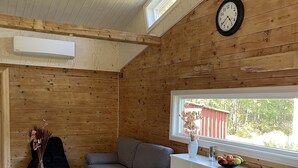 Een gezellig interieur van een chalet, met een grijze bank, een kleine witte tafel met een vaas, een wit kastje met fruit, en een houten plafond met een klok.