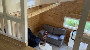 Een gezellig chalet interieur met een houten plafond, een grijze bank, een kleine witte tafel, en een raam met uitzicht op groen.