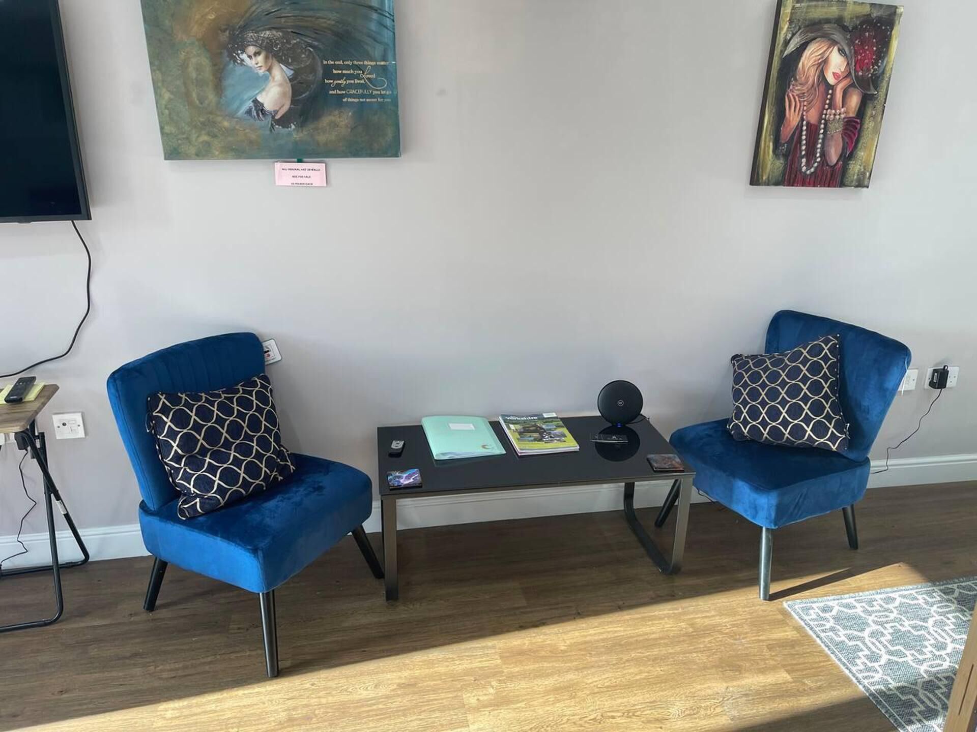 Una habitación con dos sillones azules, una mesita con un libro y un altavoz, y obras de arte en la pared.