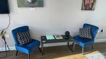 Una habitación con dos sillones azules, una mesita con un libro y un altavoz, y obras de arte en la pared.