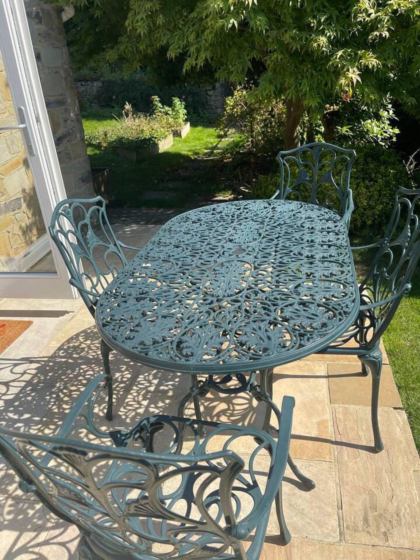 Une table d’extérieur en métal, ornée de motifs complexes, entourée de chaises assorties, placée sur une terrasse avec un mur en pierre et de la verdure en arrière-plan.