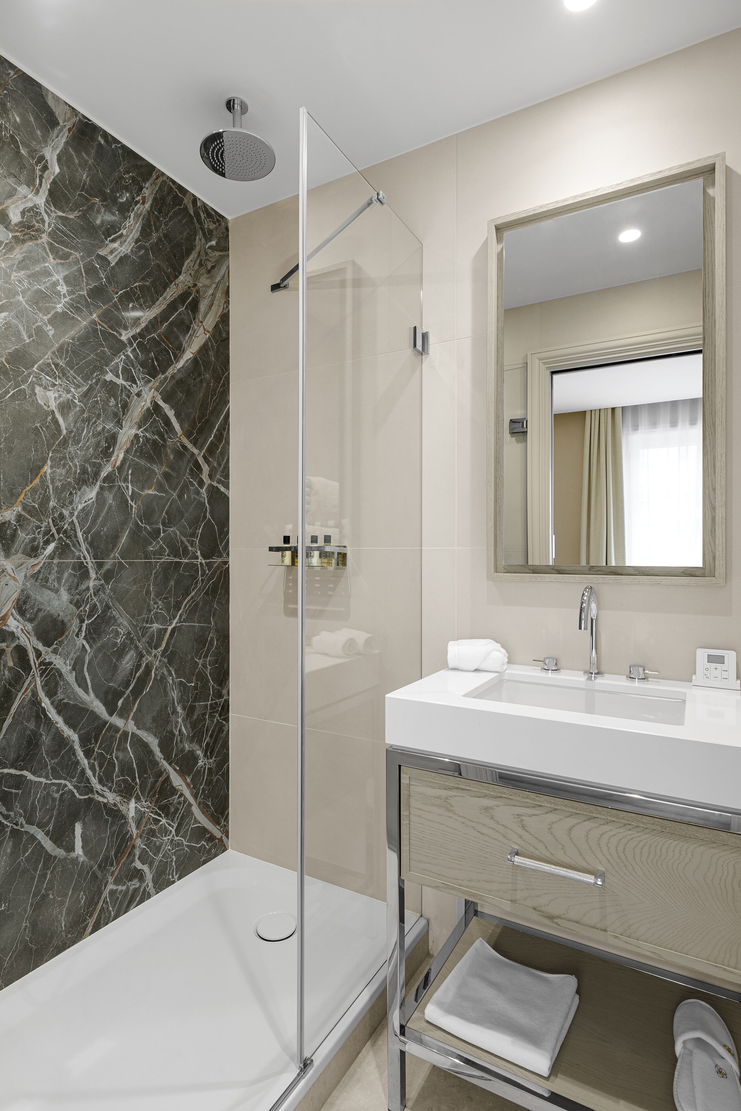 A modern bathroom with a glass-enclosed shower, a marble wall, a white sink, a mirror, and a wooden vanity.